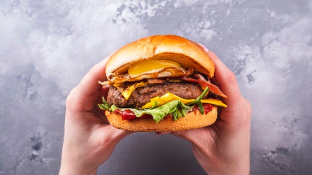 Manos femeninas sosteniendo sabrosas hamburguesas de ternera con huevo frito y tocino Vista superior Comida americana