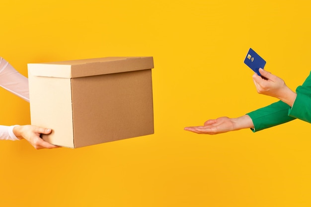 Foto manos femeninas sosteniendo tarjeta de crédito y tomando caja de cartón del transportista o vendedor en estudio amarillo