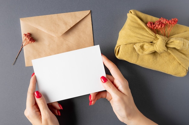 Manos femeninas sosteniendo una tarjeta de papel en blanco sobre una mesa gris con un sobre de artesanía regalo furoshiki