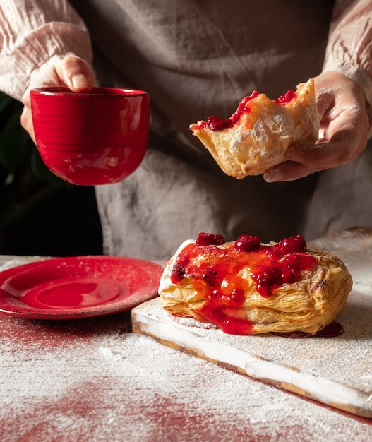 Manos femeninas sosteniendo una taza de café roja y una rebanada de hojaldre con mermelada de ciruela o grosella roja sobre la mesa.