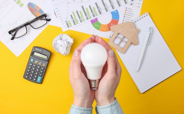 Manos femeninas sostienen una bombilla de ahorro de energía en un amarillo con gráficos, tablas y una calculadora