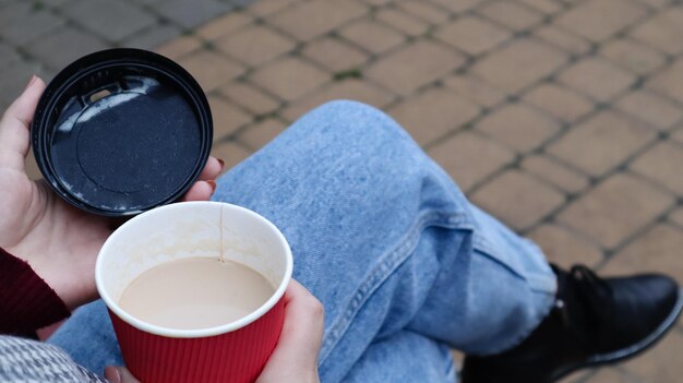 Manos femeninas sostienen café para llevar por la mañana y cierran un vaso de papel con tapa mientras están sentados en un banco del parque, primer plano de las manos.
