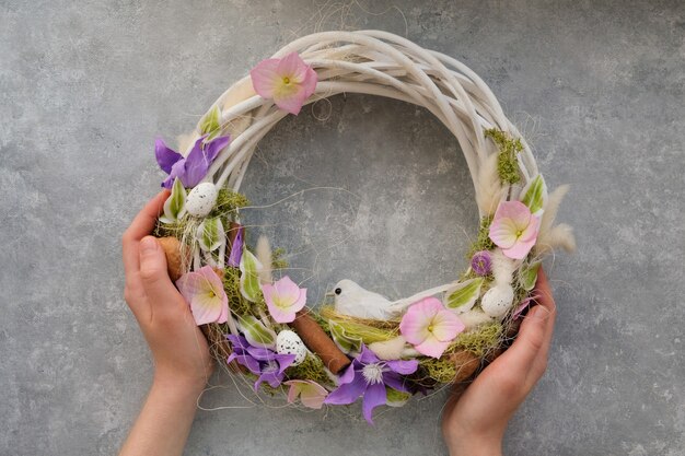 Foto manos femeninas sostienen una corona de pascua con flores y un pájaro sobre una mesa gris. concepto de primavera de decoración de la casa.