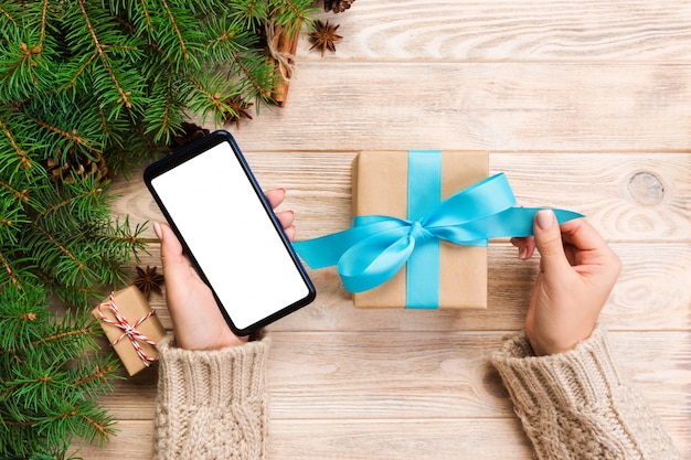 Las manos femeninas sostienen envolviendo la caja de regalo de Navidad y leyendo el masaje en el teléfono con cinta azul en la mesa de madera marrón. Vista superior