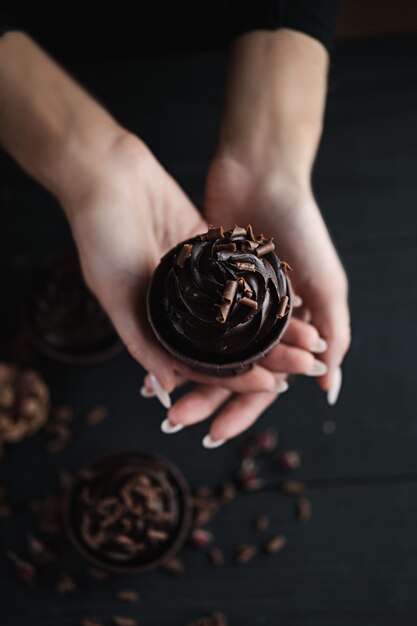 Las manos femeninas sostienen un muffin de chocolate o una magdalena sobre un fondo oscuro. Varias magdalenas o cupcakes con crema en forma de chocolate en la mesa negra. Vela festiva arde en un pastel de chocolate.