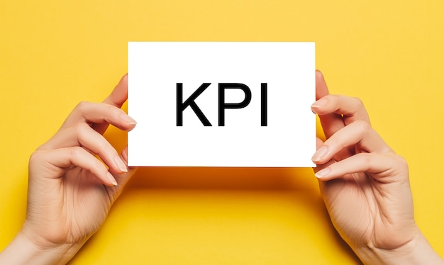 Manos femeninas sostienen papel de tarjeta con KPI de texto sobre un fondo amarillo. Concepto de negocios y finanzas