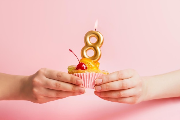 Las manos femeninas sostienen un pastel dulce con una cereza de cóctel y una vela ocho sobre un fondo rosa