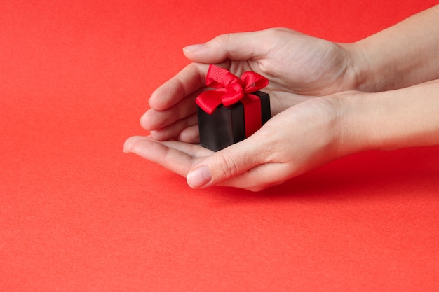 Manos femeninas sostienen una pequeña caja de regalo negra con un lazo brillante sobre un fondo rojo.