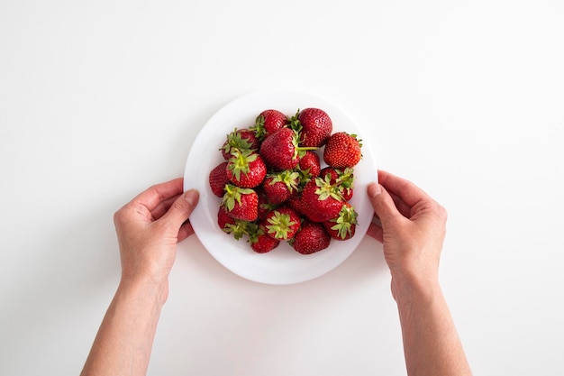 Manos femeninas sostienen un plato con fresas sobre un fondo de mesa blanca Vista superior plana