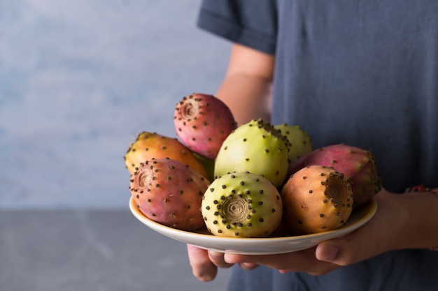 Manos femeninas sostienen un plato con frutas frescas maduras de nopal sobre un fondo gris