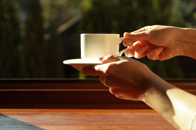 Las manos femeninas sostienen una taza de bebida caliente cerca del alféizar de la ventana