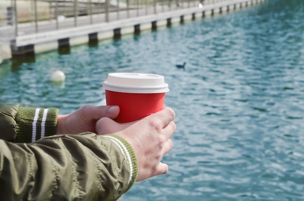 Manos femeninas sostienen una taza de cartón con café en el muelle del mar.