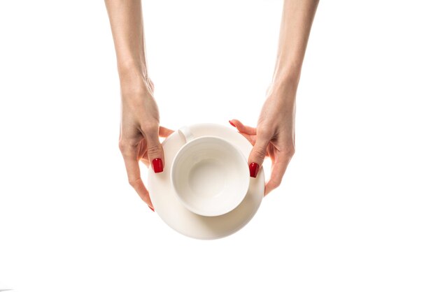 Manos femeninas sostienen una taza de cerámica blanca con un platillo sobre fondo blanco Manos femeninas con manicura roja fresca Aisladas sobre fondo blanco