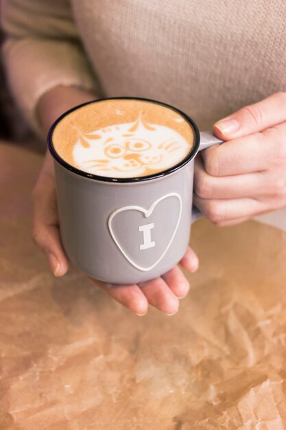 Las manos femeninas sostienen una taza esmaltada gris cálida con un símbolo de corazón.