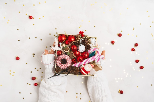 Manos femeninas en suéter de punto blanco sosteniendo una caja con accesorios de árbol de Navidad vintage