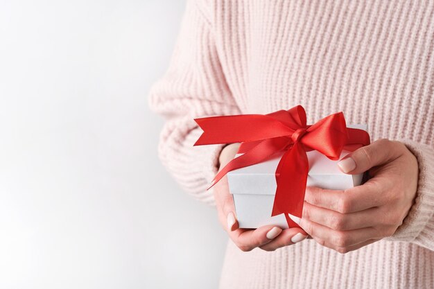 Manos femeninas en suéter rosa de lana con caja de regalo blanca con cinta roja sobre fondo claro Cumpleaños de Navidad o composición de boda con espacio de copia Mockup para Año Nuevo o Navidad