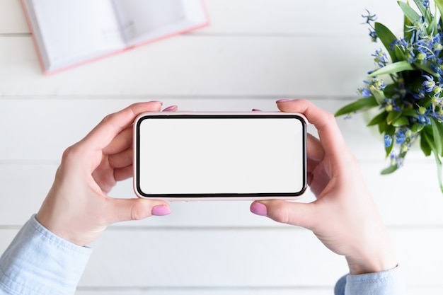 Manos femeninas con un teléfono inteligente. Pantalla blanca en blanco. Mesa con cuaderno y flores