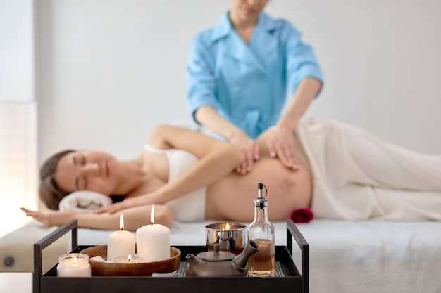 Manos femeninas del terapeuta de masaje haciendo un ligero masaje en el vientre de la mujer embarazada en la sala de cosmetología, spa o centro de bienestar en el interior. Vista lateral. centrarse en velas en la mesa