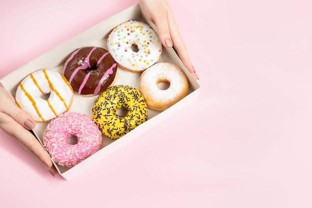 Manos femeninas tomando coloridos donuts acristalados redondos con chispitas de caja de donas en superficie rosa pastel