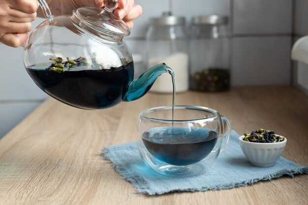 Manos femeninas vertiendo anchon de una tetera de vidrio en una taza en la cocina té de flor de guisante de mariposa azul
