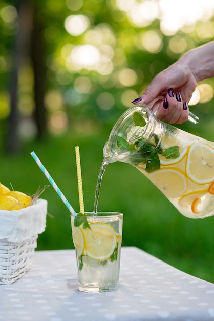 Las manos femeninas vierten refrescante limonada fría con limones en hielo y menta de una jarra en un vaso en una mesa de jardín de verano en un picnic. Enfoque selectivo suave.