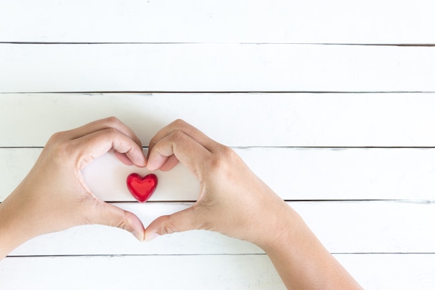 Las manos forman corazón firman sobre un corazón rojo en el fondo de madera blanco