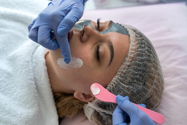 Las manos con guantes azules aplican crema hidratante blanca en la cara del paciente para el cuidado de la piel. Concepto de piel corporal limpia y suave