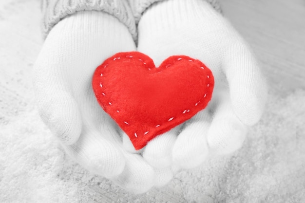 Foto manos en guantes blancos cálidos con corazón rojo sobre fondo nevado