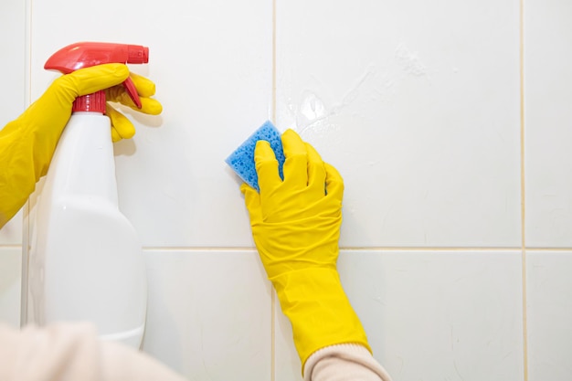 Manos con guantes de goma amarillos lavando los azulejos en el baño. De cerca.