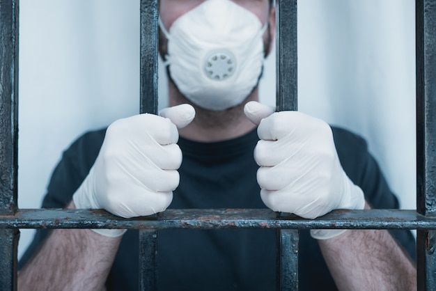 Manos con guantes médicos en barras. Hombre aislado con máscara de cirugía agarra las barras de la ventana