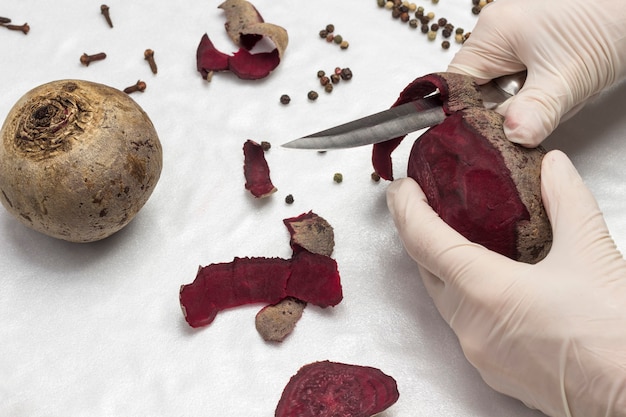 Las manos en guantes pelan la remolacha. Alimentos saludables para el invierno. Superficie blanca. Vista superior