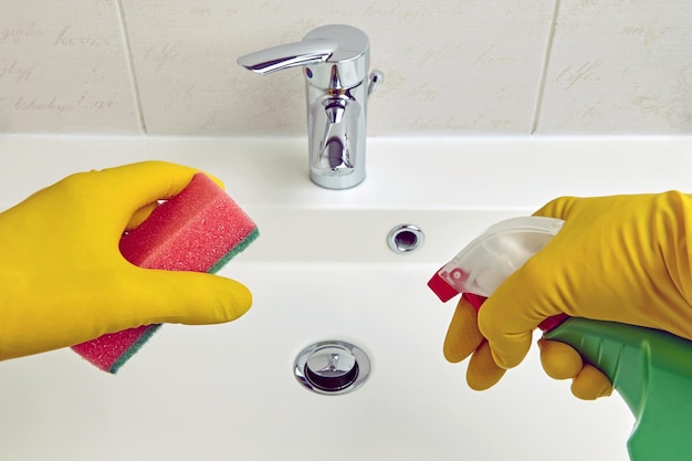 Las manos con guantes protectores amarillos sostienen una esponja y un agente de limpieza para limpiar equipos sanitarios en el baño