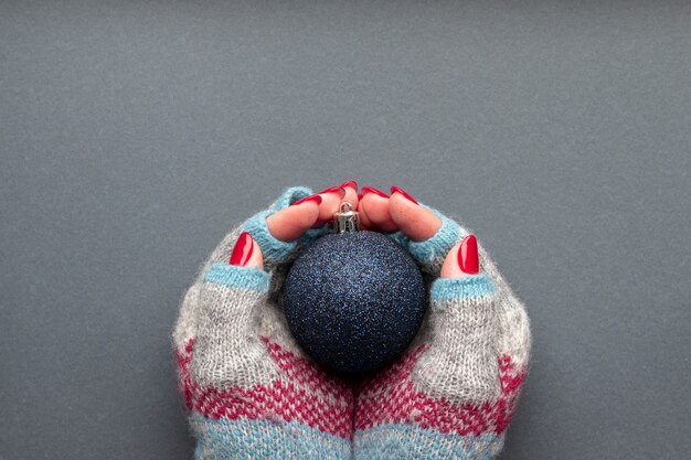 Manos en guantes tejidos con bola de Navidad brillante y con uñas rojas brillantes sobre gris