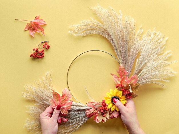 Manos haciendo corona floral seca de hierba de pampa seca y hojas de otoño Yacía plana sobre papel amarillo luz del sol con largas sombras