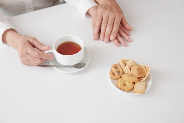 Manos de hijo adulto tocando la mano de su mujer madura cuando están bebiendo té con galletas en casa