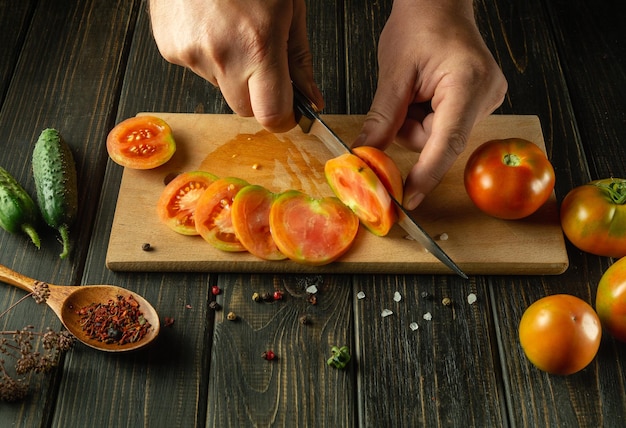 Las manos de un hombre con un cuchillo cortando un tomate en una tabla de cortar El concepto de cocinar ensalada de verduras con pepinos y tomates en casa