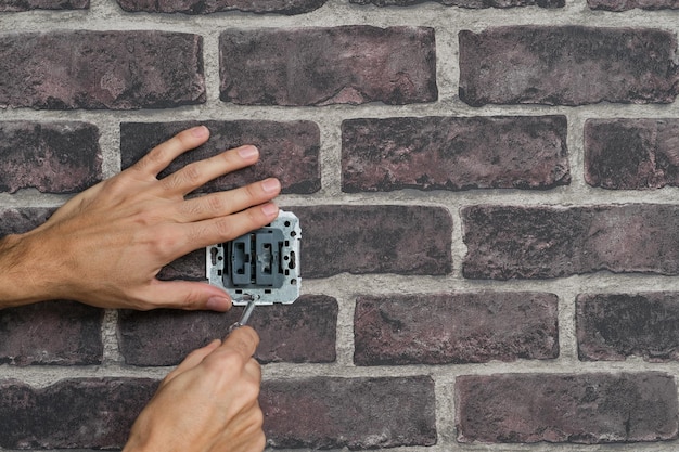 Foto manos de hombre con destornillador desmontando un interruptor instalado en una pared de ladrillo falso