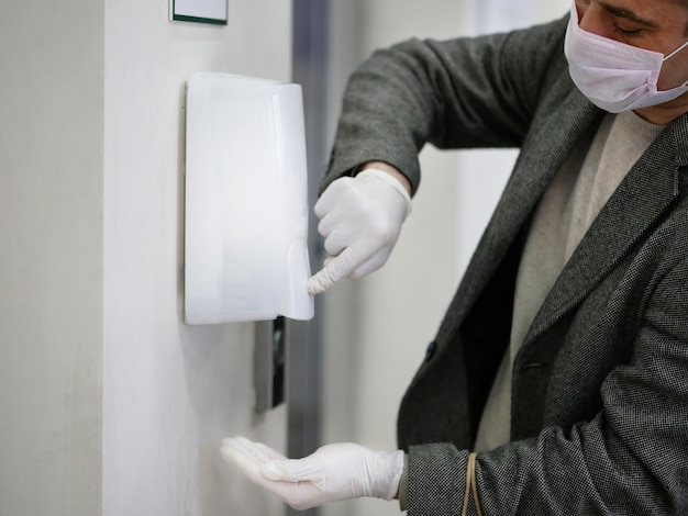 Manos de hombre en guantes usando dispensador automático de alcohol para limpiar bacterias y virus corona