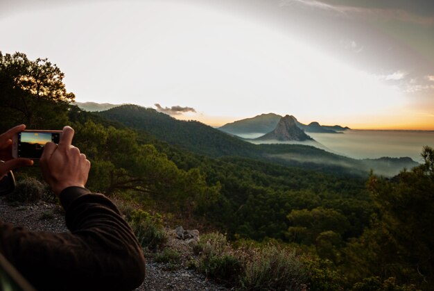 Manos del hombre haciendo fotos en su teléfono de la hermosa vista a la montaña y al mar en la niebla y el atardecer Chipre