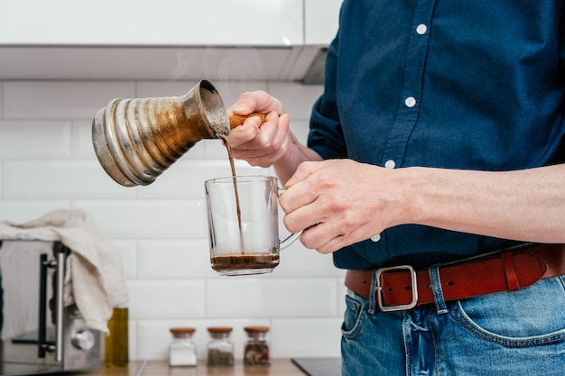 Manos de un hombre irreconocible con camisa azul que vierte café recién hecho de cezve de cobre en una taza de vidrio en una cocina brillante