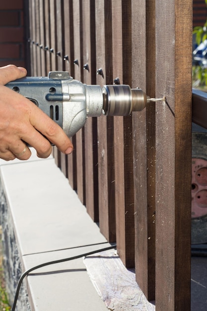 Manos del hombre perforando una cerca de madera para una construcción metálica Construyendo una cerca de madera con un taladro y un tornillo Cerca de su mano y la herramienta en un concepto de bricolaje