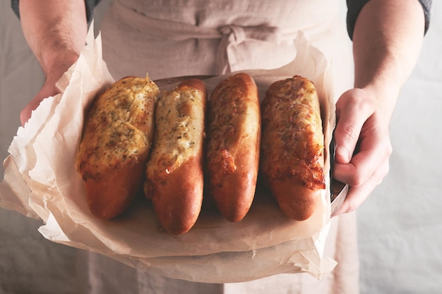 Manos de hombre sosteniendo sándwich horneado caliente en pan baguette con jamón, tocino, verduras y queso en pergamino Hombre en delantal negro en el fondo de la cocina casera con panadería Maqueta