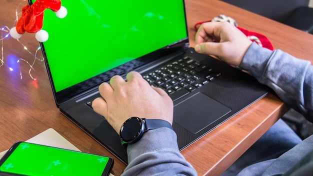 manos de hombre y teclado de portátil con pantalla verde vacía maqueta de teléfono inteligente y decoración de Navidad