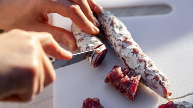 Manos de hombres cortadas con cuchillo salchicha curada francesa con moho blanco en el primer plano de la tabla de cortar