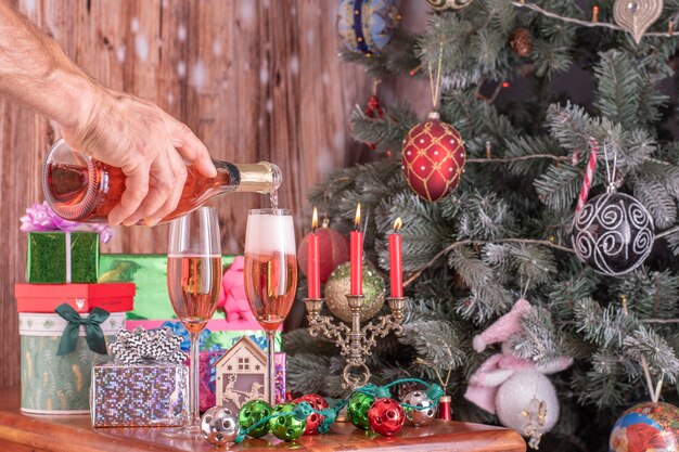 Las manos de los hombres vertiendo champán de una botella en flautines de champán con el telón de fondo de un árbol de año nuevo y regalos.