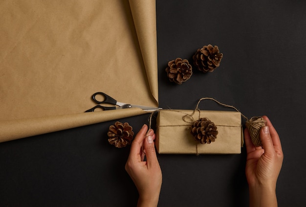 Manos humanas sosteniendo un presente rústico simple en papel de regalo artesanal decorado con una piña y un lazo atado con una cuerda. Mujer decorando regalos de Navidad. Preparación navideña.