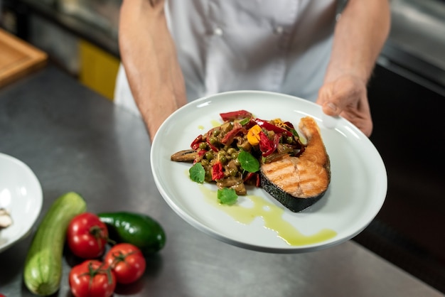 Foto manos del joven chef profesional sosteniendo un plato con un trozo de salmón frito y verduras picadas al vapor en la cocina
