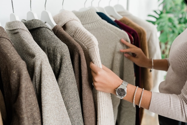 Manos de joven comprador en jersey gris mirando a través de la nueva colección de temporada de ropa casual en boutique