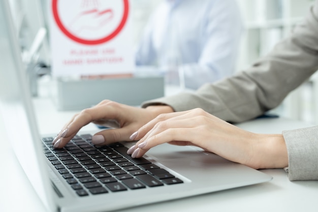 Manos de joven empresaria contemporánea en ropa formal presionando las teclas del teclado de la computadora portátil durante la red mientras está sentado en el escritorio en la oficina