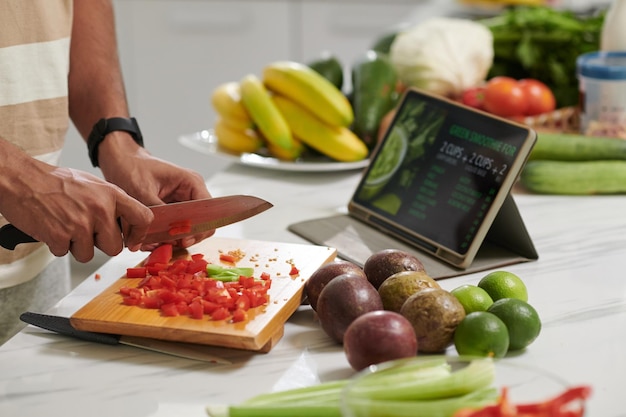 Foto las manos de un joven moderno con un cuchillo afilado cortando capsicum rojo fresco en una tabla de madera mientras sigue los puntos de la receta en línea